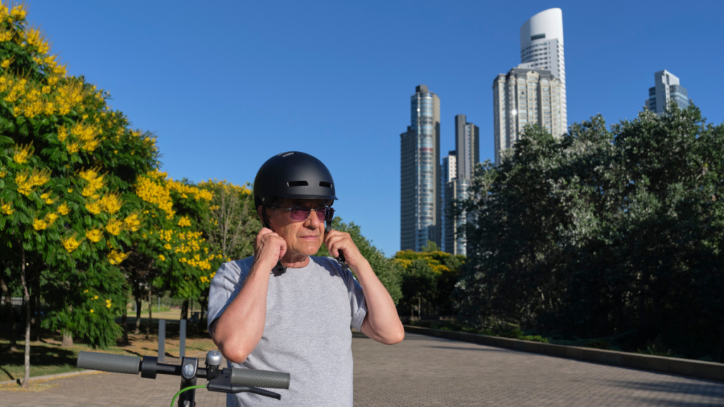 Muškarac u kratkoj sivoj majici stavlja crnu kacigu na glavu dok stoji pored električnog romobila