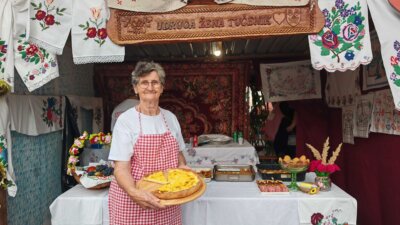 Katarina Bernardić u bijeloj majici i cvrnoj kariranoj pregači drži u ruci tanjur s jelom 'vrboavačko perje'