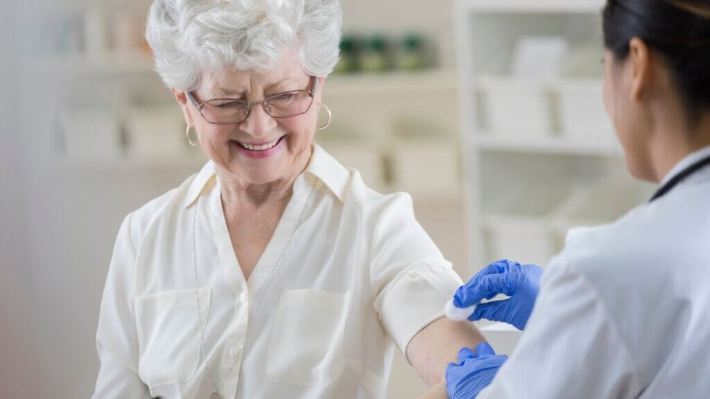 Umirovljenica kratke sijede kose s naočalama u bijeloj košulji kratkih rukava pruža ruku liječnici koja briše ruku vaticom prije cijepljenja