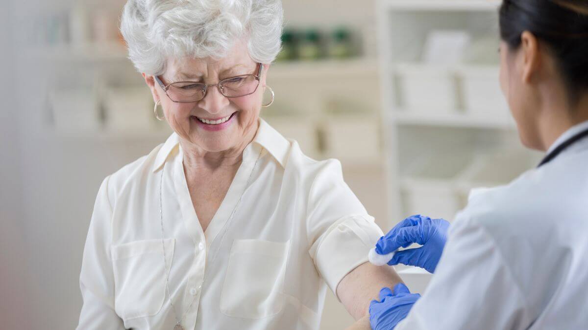 Umirovljenica kratke sijede kose s naočalama u bijeloj košulji kratkih rukava pruža ruku liječnici koja briše ruku vaticom prije cijepljenja