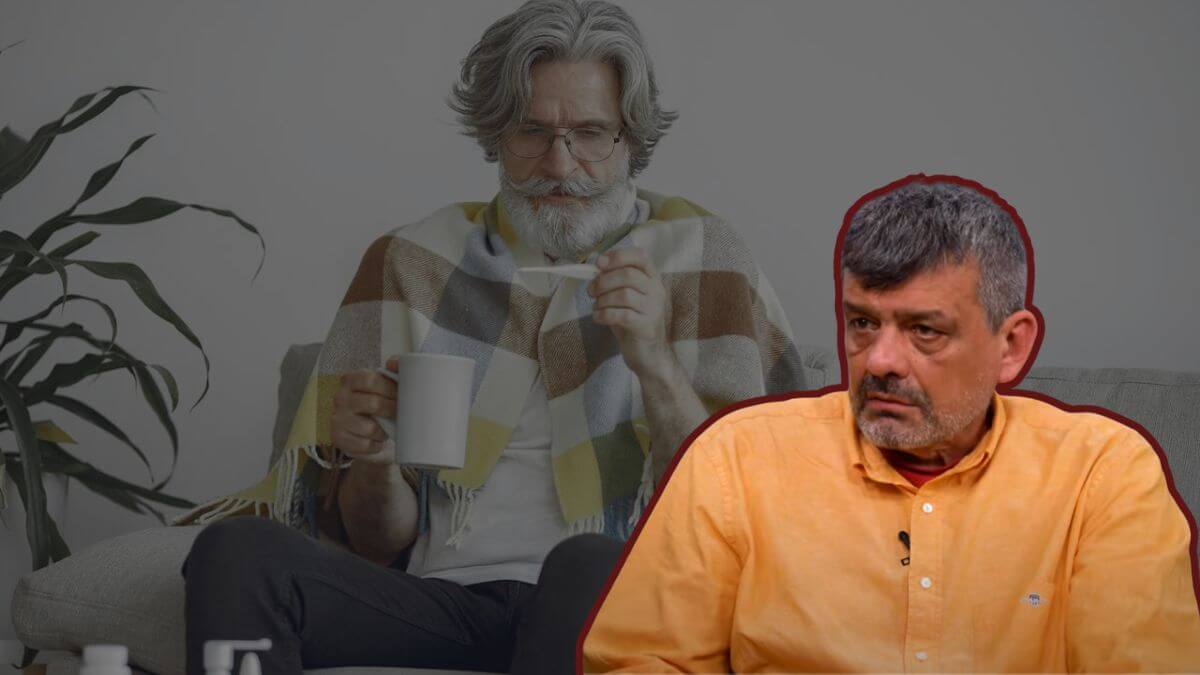 Stariji muškarac na kauču obgrljen dekom, u jednoj ruci šalica, u drugoj gleda u toplomjer, pored njega epidemiolog Bernard Kaić u narančastoj košulji