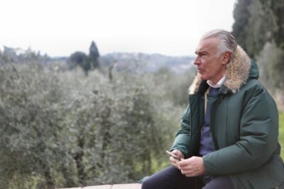 Stariji muškarac sijede kose sjedi vani na ogradi u zelenoj jakni s krznenom kapuljačom, iza njega drveće