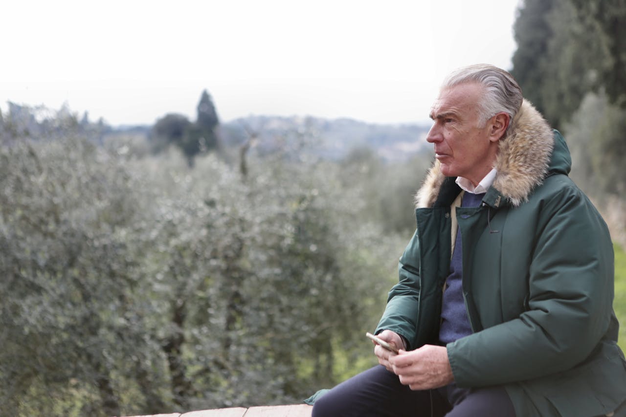 Stariji muškarac sijede kose sjedi vani na ogradi u zelenoj jakni s krznenom kapuljačom, iza njega drveće