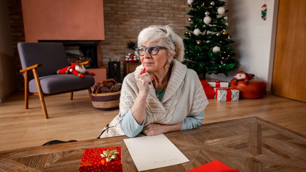 Umirovljenica sjedi na podu i piše pismo na stoliću, iza nje okićeni bor i pokloni